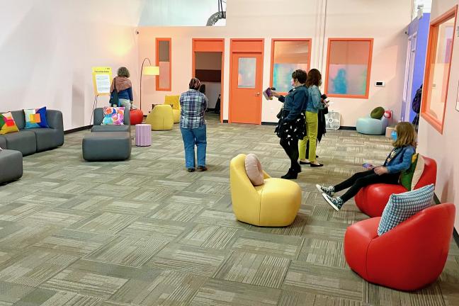 A crowd of people walks around a room with colorful walls and furniture.