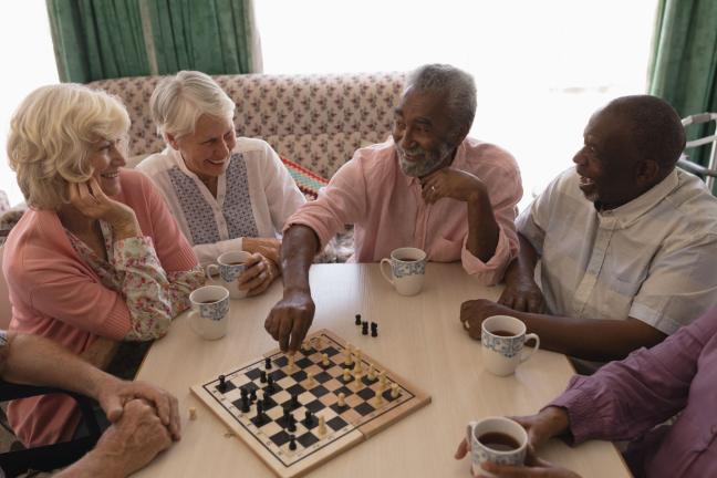 A diverse group of older adults playing chess at a table while drinking coffee and laughing