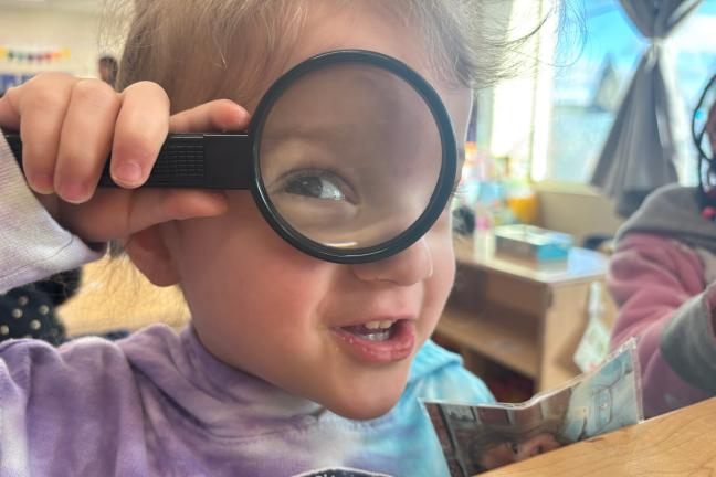 young preschool student holds up magnifying glass to camera. 