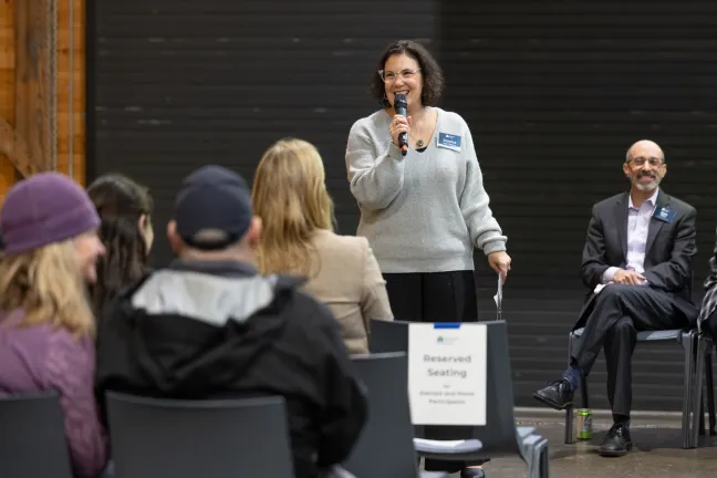 A woman (Chair Jessica Vega Pederson) speaks into a microphone at a gathering. People are seated facing her.