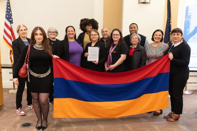 A group of people stand indoors holding a large red, blue, and orange horizontal striped flag. The American flag and the Oregon flag are visible in the background. One person holds a document.