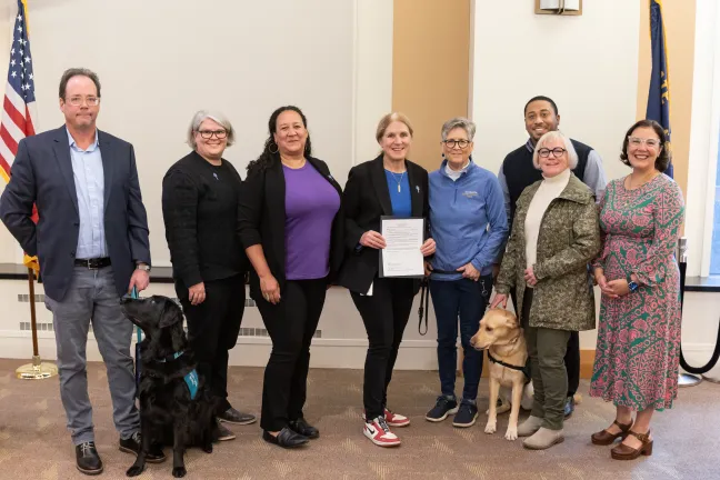 A group of eight adults and two dogs pose indoors with American and another flag. One adult holds a framed document. The setting is the Multnomah County Board Room.