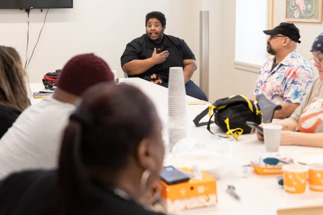 A person speaks to an audience of people seated around a white table
