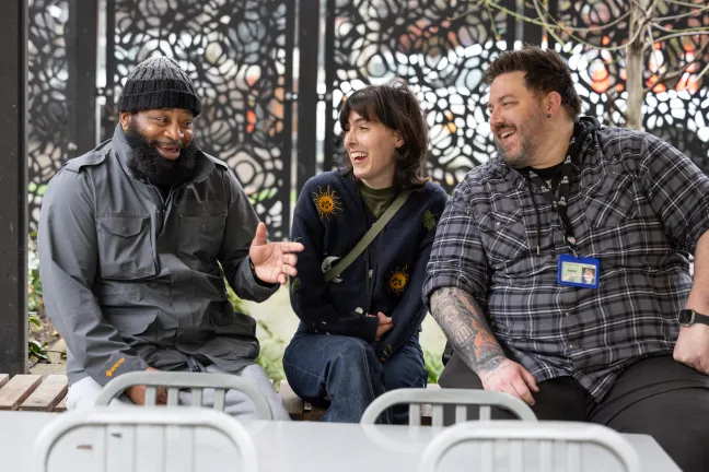 three people sitting on a bench in the Behavioral Health Resource Center courtyard smiling in conversation