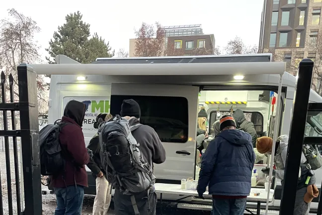 People stand in front of a folding table setup outside a white van with its doors open.