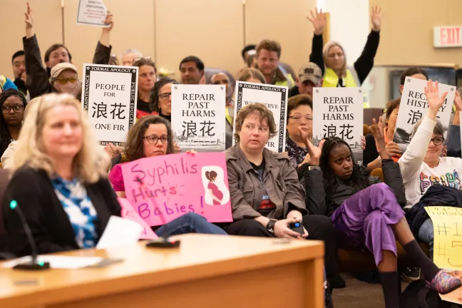 Community members sit and stand at budget hearing. Many are holding signs.
