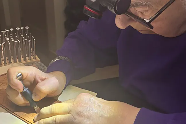 A close-up shot of David Cohen wearing a purple sweater and a head-mounted magnifier, meticulously working on a piece of light brown leather with a hand-held engraving instrument.