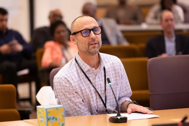 A man with a beard and glasses sits at a table, speaking into a microphone. He wears a plaid shirt and an ID badge on a lanyard. A box of tissues is to his left, and papers are in front of him. Other people are seated in the background, out of focus.