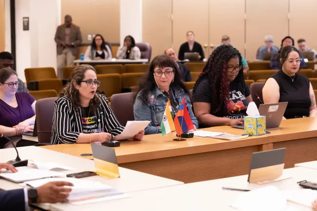 Five people are seated at desks in a Board room. One speaks while holding papers, with two small flags in front of them. Several other people are visible in the background.