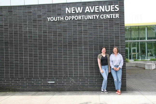 Two people stand side-by-side outdoors, smiling in front of a dark brick wall. Above them, white letters on the building read, "NEW AVENUES YOUTH OPPORTUNITY CENTER".