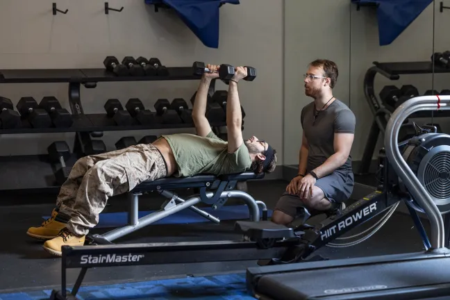 Jayson, lies on a workout bench, lifting two dumbbells above his chest. Levi sits behind him, spotting him. 