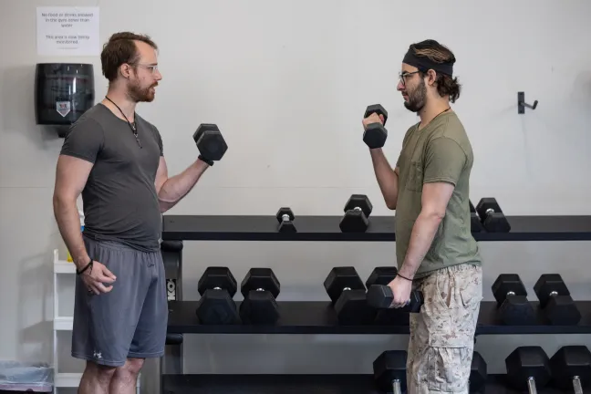  Two men stand facing each other in a gym, each holding a dumbbell.