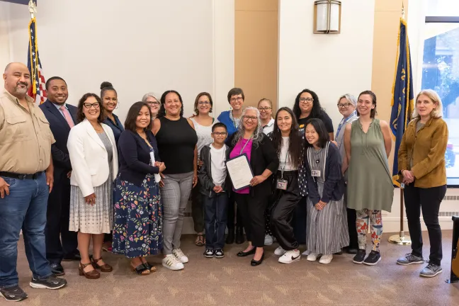 A smiling group of people pose for the camera between an American flag and a State of Oregon flag. A person toward the center of the group holds up the proclamation.