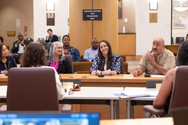 For people are sitting at the dais. The right-most person is speaking to the commissioner in the foreground. The two people on the left are looking toward the speaker; the third person from the left is looking at the commissioners.
