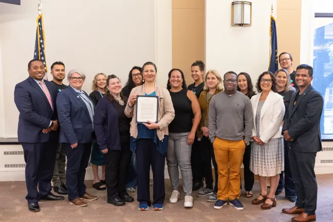 A smiling group of people pose for the camera between an American flag and a State of Oregon flag. A person toward the center of the group holds up the proclamation.