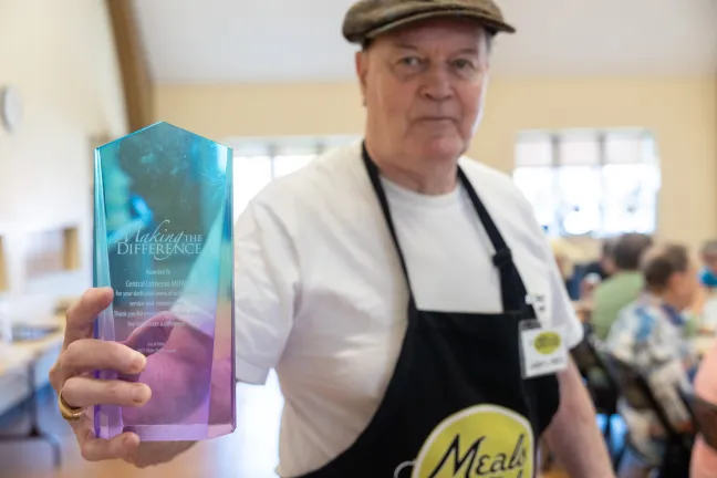 A man holds a prism-shaped, translucent award that reads "Making the Difference." The award is light blue and purple. The man is looking directly at the camera.