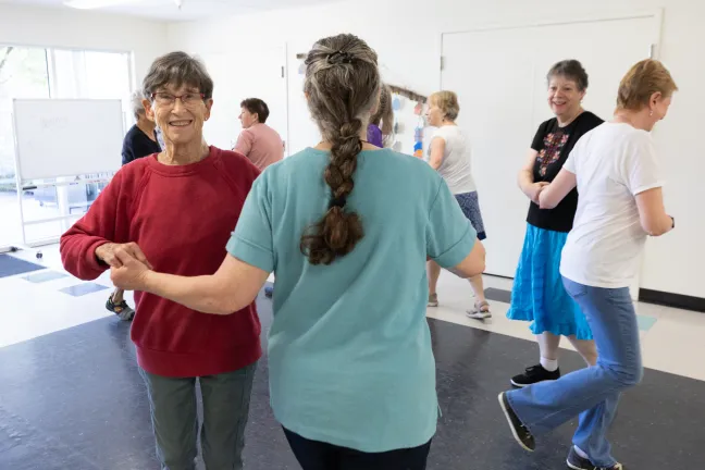 A group, dressed in casual clothing and skirts, holds hands in a circle as they participate in a folk dance class indoors.