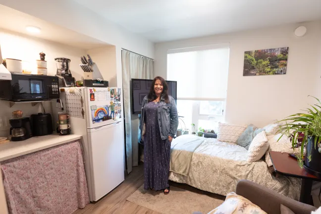Cori stands in front of her bed in a small apartment.