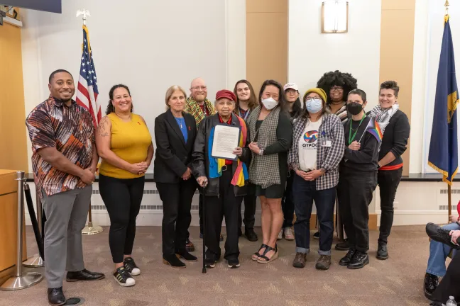 County staff and community members stand with Commissioners in the boardroom. One person in the middle holds the signed proclamation.