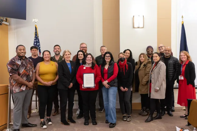 County staff and community members stand with Commissioners in the boardroom. One person in the middle holds the signed proclamation.