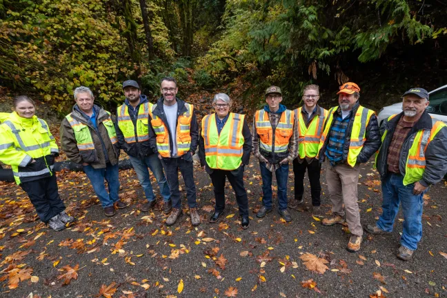 a group of smiling people posing in safety vests on a road covered in fallen leaves