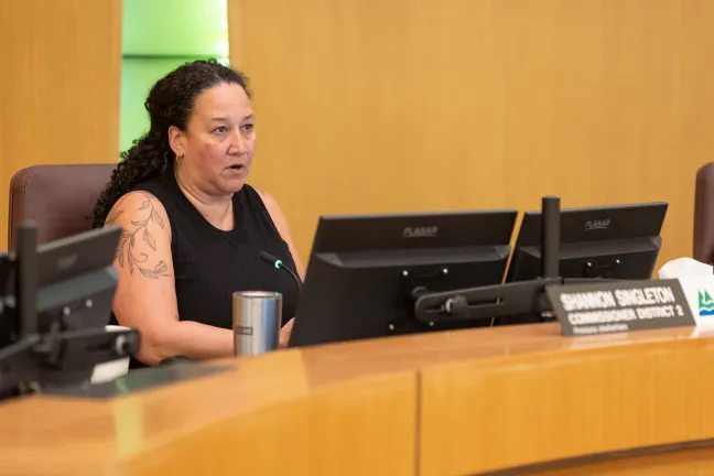 Commissioner Shannon Singleton speaking at the Multnomah County Board dais. She is seated behind a nameplate that reads "Shannon Singleton, Commissioner District 2," and several computer monitors are visible in front of her.