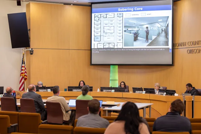 A wide shot of the Multnomah County Board of Commissioners meeting room. Five commissioners sit at a curved dais beneath a large projector screen showing "Sobering Care" facility floor plans and 3D renderings. Several people in the audience face the dais.