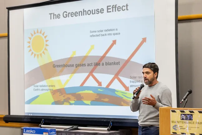 Un ponente habla sobre el efecto invernadero en una presentación sobre planificación de impactos climáticos. En la pantalla de fondo se muestra un diagrama de la atmósfera terrestre actuando como una manta que atrapa el calor.