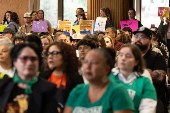  A packed room of comunity memebrs seated and standing along the back of the room, holding posters in support of Save SUN 