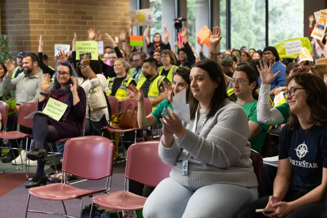 Community members sitting clapping and waving hands in support of others testifying in room. 