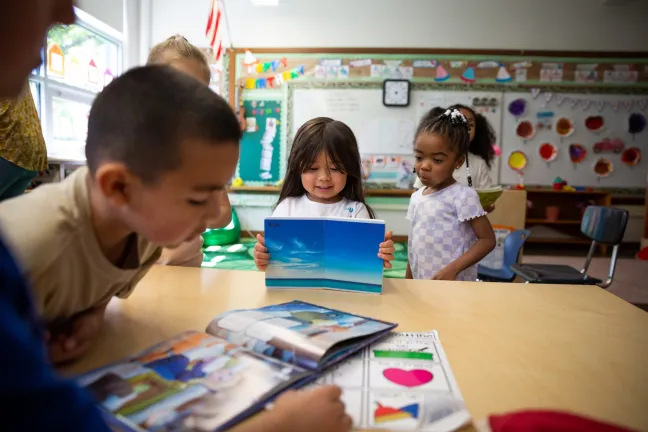 A young child smiles while holding up a picture book about the ocean for their classmates at a table inside a bright preschool classroom.