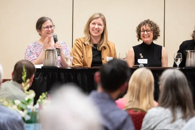 Three people are seated on a stage behind a long table with a black tablecloth, all laughing and smiling warmly. T