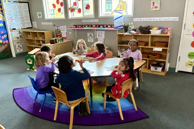 Seven preschool aged children sit around a table eating lunch in a classroom with filled with toys, books, and artwork. 