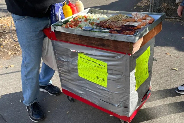 Bacon-wrapped hot dogs heating on top a makeshift food cart on a corner.