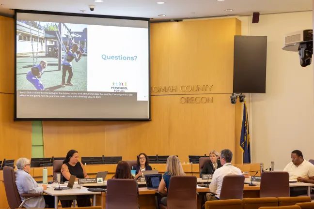 The Multnomah Board of County Commissioners and  representatives from the Preschool and Early Learning Division, sit at a large round table inside of the Multnomah County Board Room. 