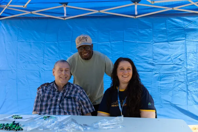 Tres personas frente a un fondo azul; dos están sentadas en sillas y la tercera está de pie detrás y entre ellas, todas sonriendo.