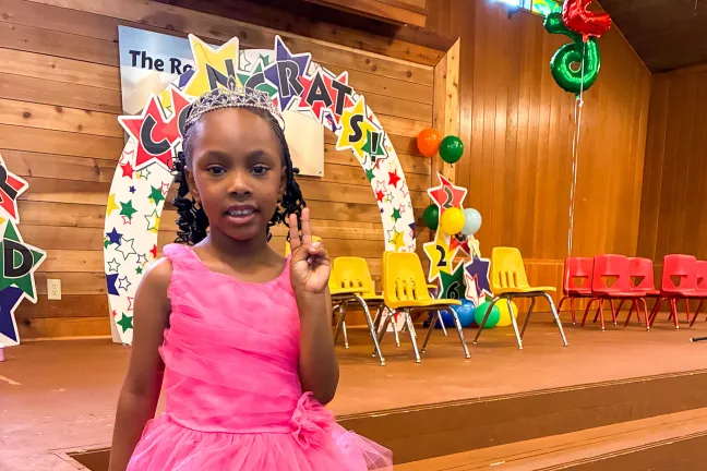 Una niña en edad preescolar luce un vestido de tul rosa y una tiara plateada mientras posa frente a un escenario con una pancarta que dice "¡Felicidades!".
