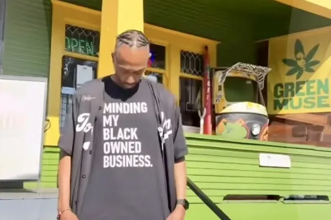Un hombre está de pie frente a un edificio verde y amarillo. Al fondo se ve un letrero que dice "Green Muse". El hombre mira fijamente su camiseta, que lleva la inscripción "Cuidando mi negocio de propiedad afroamericana".