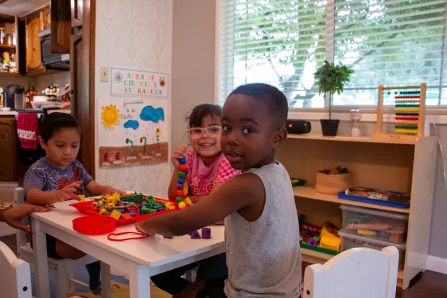 Several preschool children sit at tables working on projects in their classroom as their teacher squats down to assist them. 