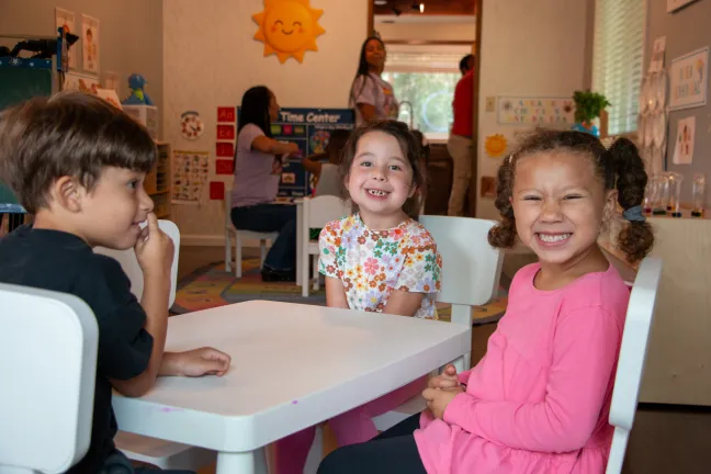 Preschool children sit at a  table in their classroom, smiling at the camera.