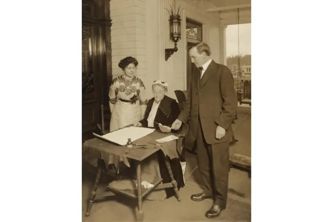 Fotografía histórica de Abigail Scott Duniway firmando la Proclamación de Sufragio Igualitario de Oregón el 30 de noviembre de 1912, mientras el gobernador Oswald West y Viola M. Coe observan.