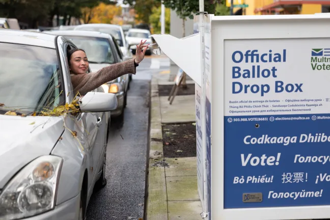 A person dropping a ballot into an Official Ballot Drop Box from a car window.