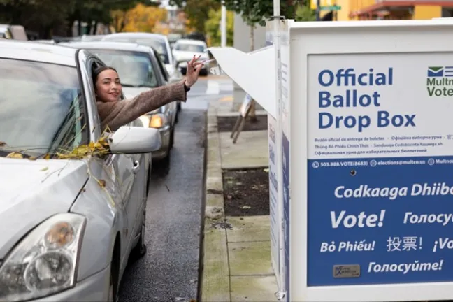 A person dropping a ballot into an Official Ballot Drop Box from a car window.