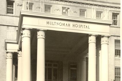 Front portico entrance of multi-story hospital building with someone standing in the open doorway/ Front of portico reads Multnomah Hospital.