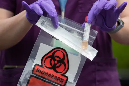 close up of a health worker in purple scrubs dropping a specimen vial into a red biohazard bag