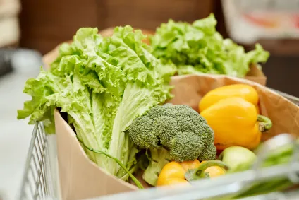 Brown paper bags filled with fresh lettuce, broccoli, and yellow bell peppers sitting inside a grocery cart.