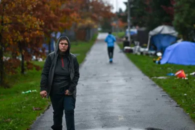 Person in gray pants and hoodie, walking towards camera on the I-205 bike path in Southeast Portland; a jogger with a blue windbreaker jogs away from camera.  Tents line the trail. Photo credit: Justin Yau