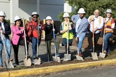 A group of people wearing hard hats hold shovels and break ground on the Arbor Lodge shelter.