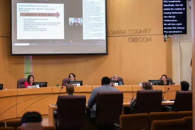Photo shows County commissioners sitting at the dias with a screen above them showing presentation slides.