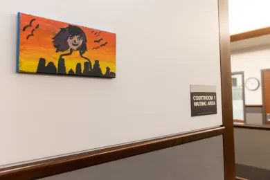 A painting of a smiling figure against a background fading top to bottom from bright orange to yellow hanging on a wall next to a sign that says Courtroom 1 Waiting Area